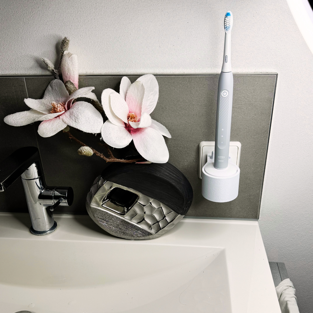 Oral B Pulsonic Slim Clean Zahnbürstenhalterung die direkt in eine Steckdose eingesteckt ist.Die Halterung ist so gestaltet, dass keine Kabel sichtbar sind. Praktische Lösung, um die Zahnbürste aufzubewahren und gleichzeitig den Raum ordentlich zu halten.